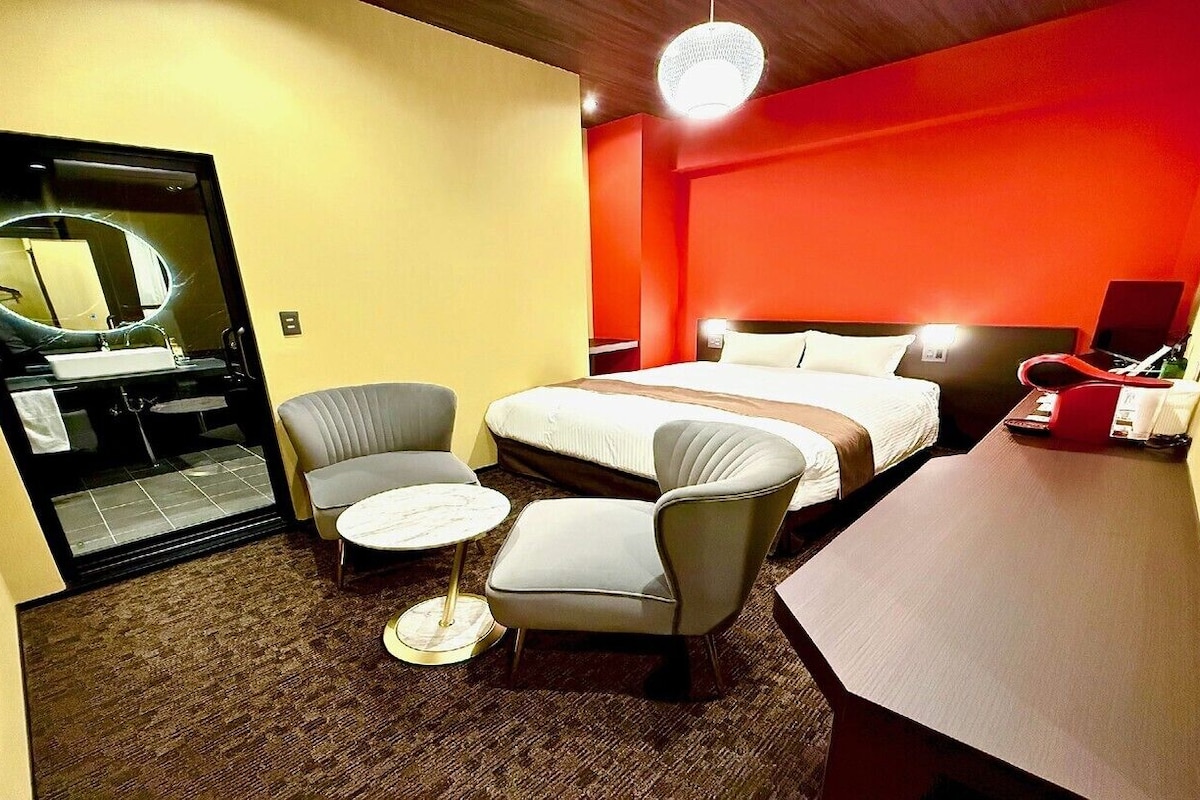 Habitación de hotel con cama, dos sillones, una mesita redonda, un escritorio, un espejo y una pared de acento roja.