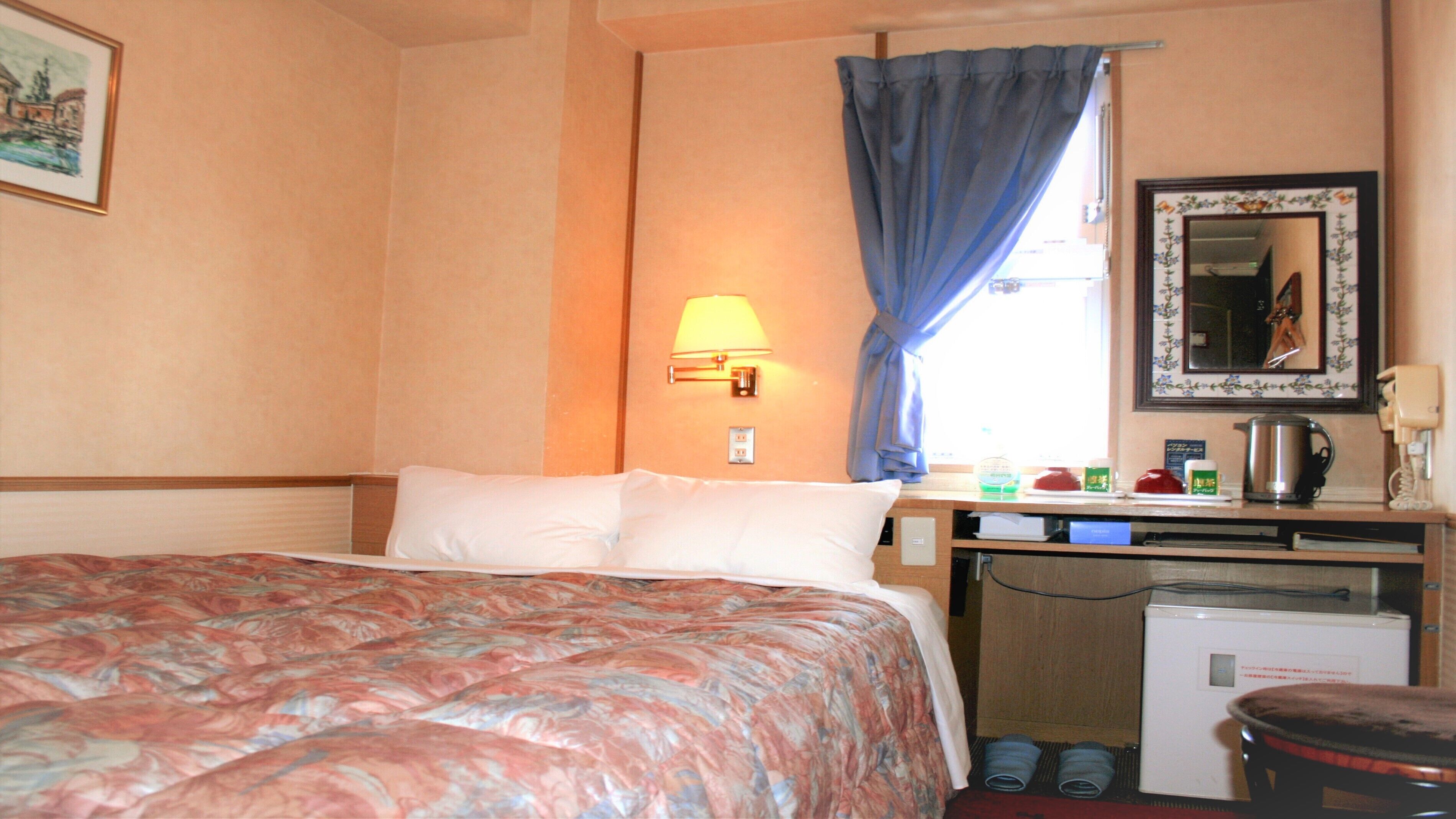 Quarto de hotel com cama, escrivaninha com luminária, espelho e cozinha compacta.