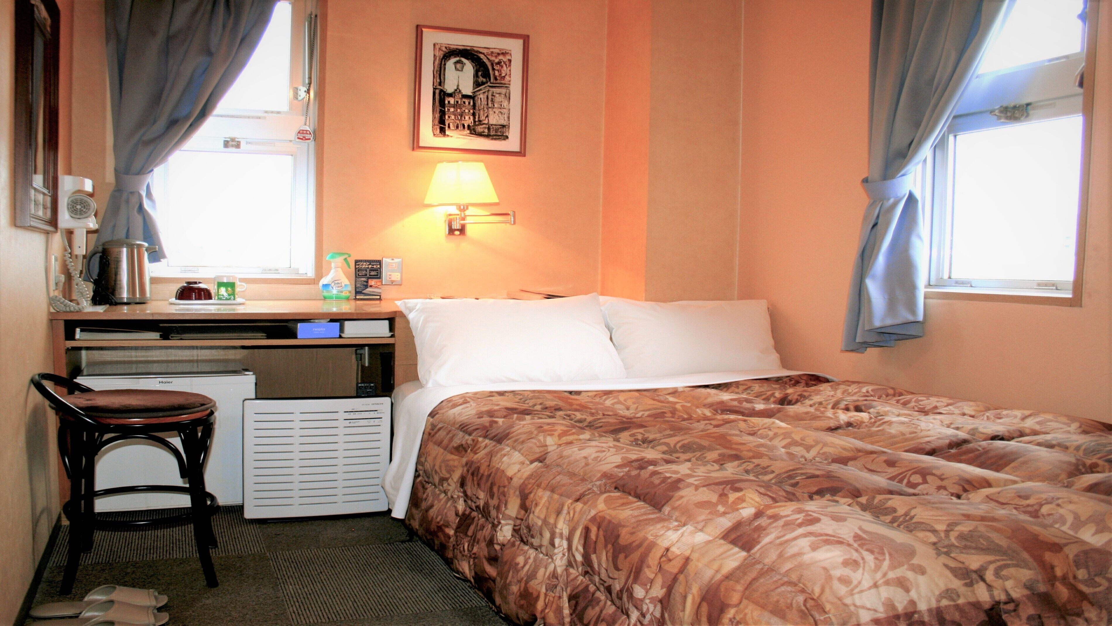 Quarto de hotel com cama, escrivaninha com cadeira, aquecedor, janela com cortinas e luminária fixada na parede.