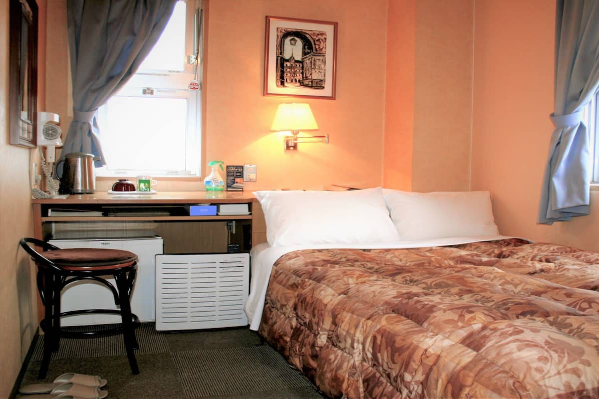 Une chambre d’hôtel comprenant un lit, un bureau avec une chaise, un radiateur, une fenêtre avec des rideaux et une lampe fixée au mur.