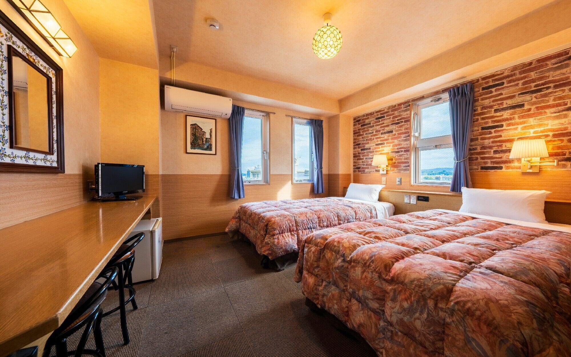 Quarto de hotel com duas camas, uma escrivaninha com televisão, uma janela com cortinas, uma parede de tijolos e uma luminária no teto.