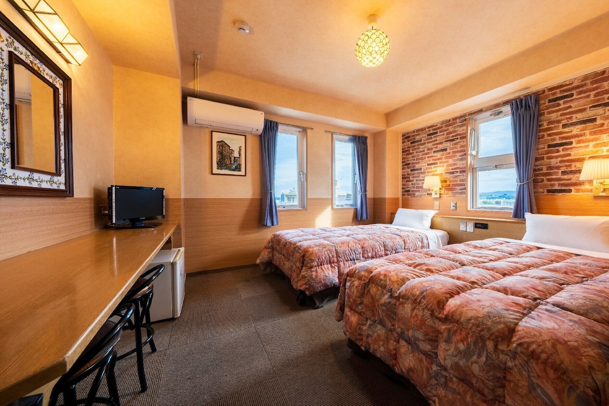 Une chambre d’hôtel avec deux lits, un bureau avec une télévision, une fenêtre avec des rideaux, un mur en briques et un luminaire au plafond.