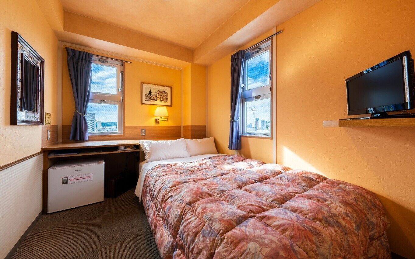 Quarto de hotel com cama, escrivaninha, televisão e janela com cortinas.