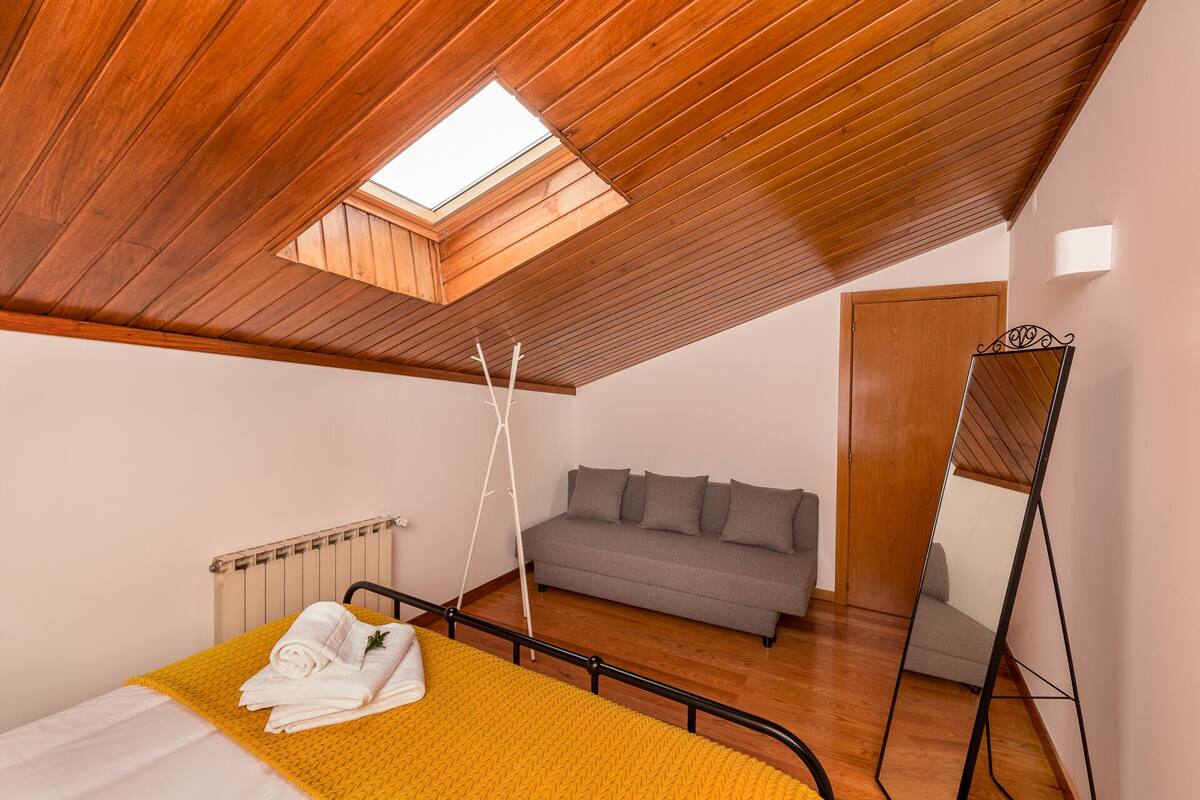 Une chambre avec un plafond en bois, une lucarne, un lit recouvert d’une couverture jaune, un canapé gris et un grand miroir.