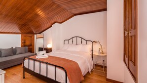 A bedroom with a sloped ceiling, a bed with a metal frame, a bedside table with a lamp, a gray sofa, and a wooden door. - Fonte Branca Guest House (Braga)