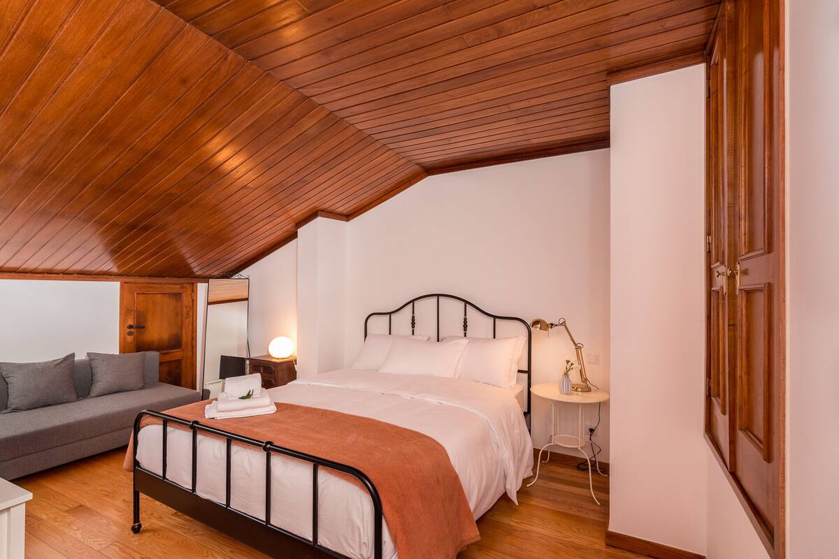 Une chambre avec un plafond en pente, un lit avec un cadre en métal, une table de chevet avec une lampe, un canapé gris et une porte en bois.