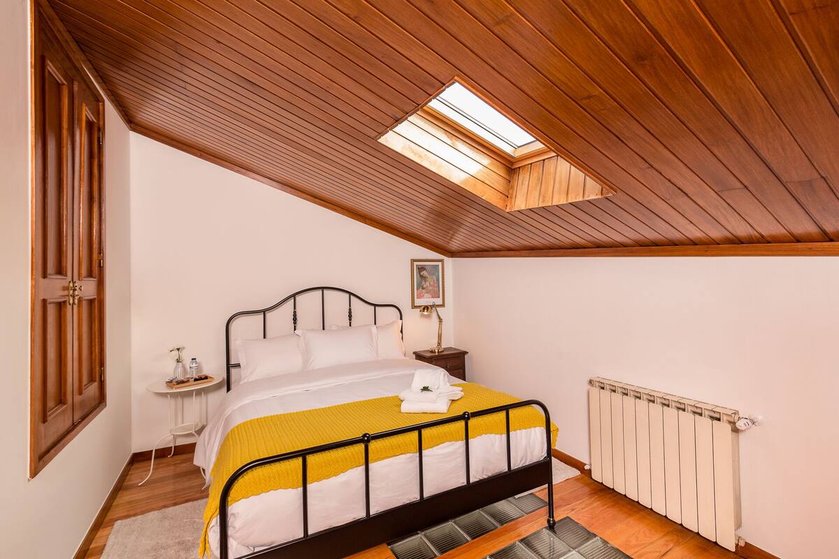 Une chambre avec un plafond en pente, un puits de lumière, un lit recouvert d’une couvre-lit jaune et blanche, une table de chevet avec une lampe et un radiateur.