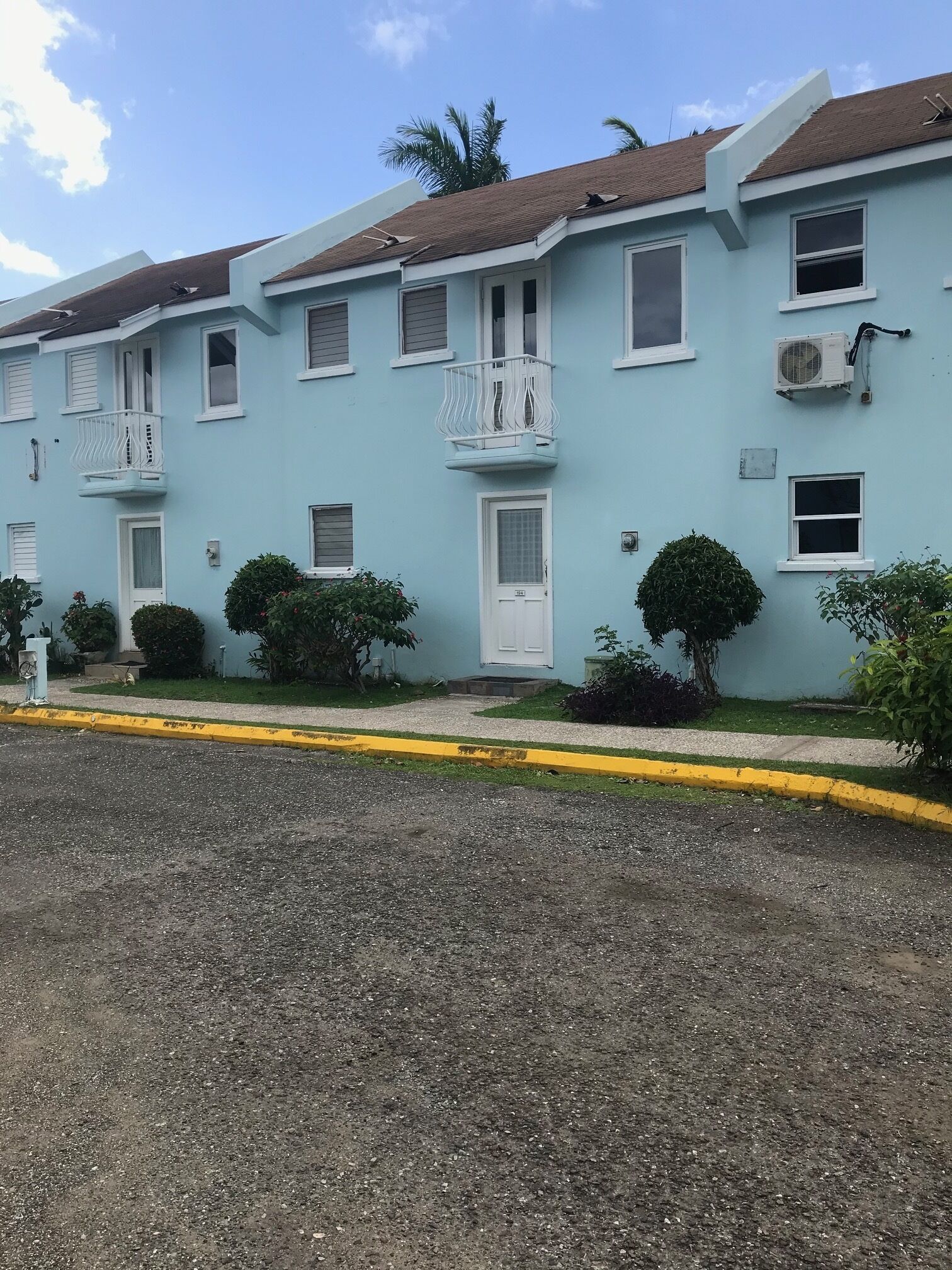 A row of light blue buildings with white balconies, a gravel driveway, and small bushes.