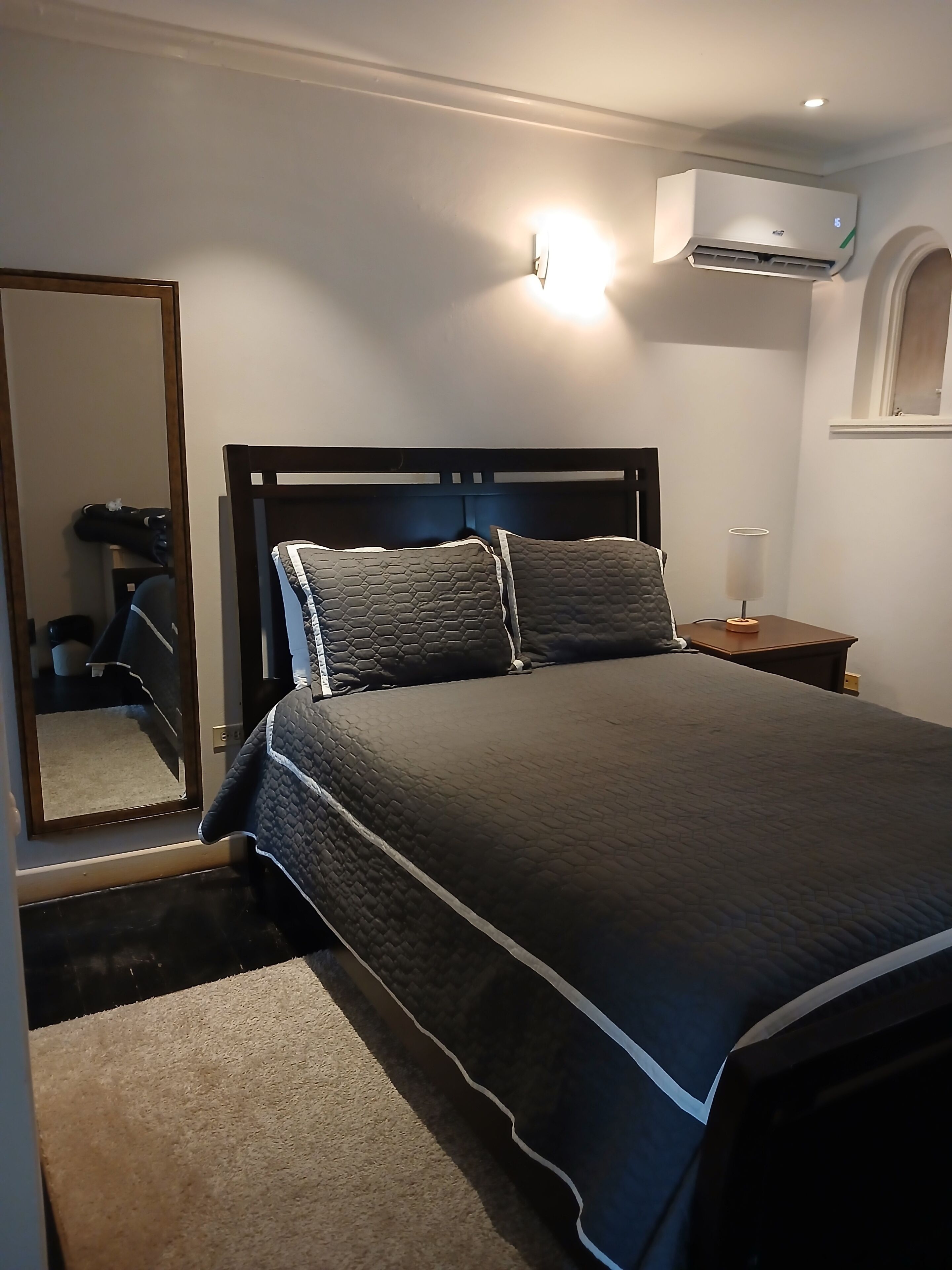 A bedroom with a bed, a nightstand, an air conditioning unit, a mirror, and a door.