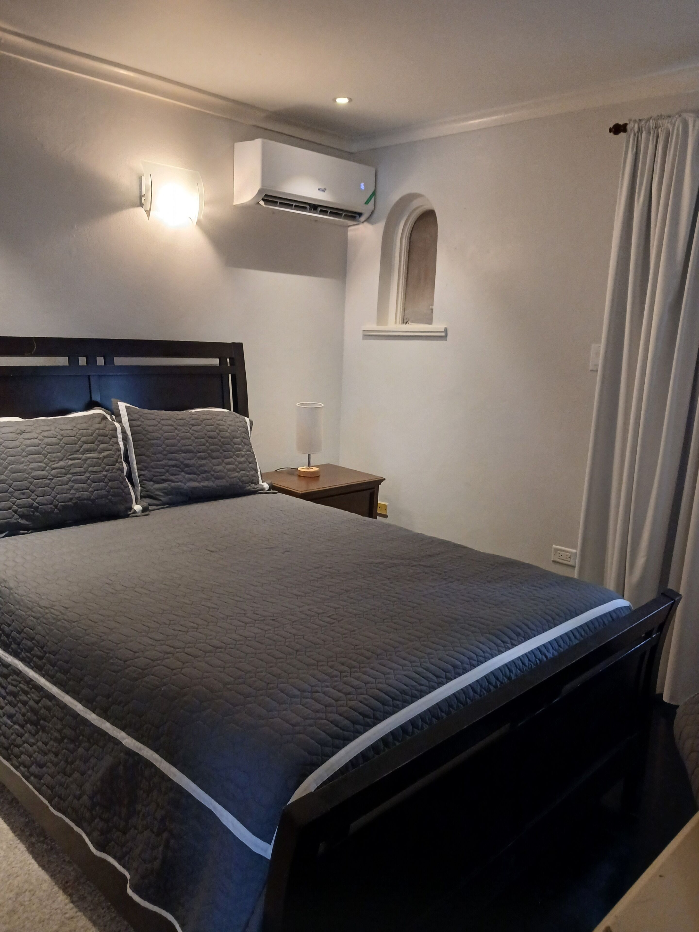 A neatly made bed with a dark quilt, a bedside table with a lamp, an air conditioning unit, and a window with curtains.