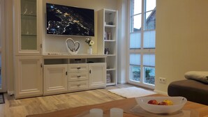 Un salon moderne avec un téléviseur à écran plat fixé au mur, un meuble blanc avec des tiroirs, une petite table sur laquelle se trouve un saladier rempli de fruits, et une fenêtre équipée de stores.