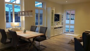 Un coin repas avec une table en bois, des chaises et un lustre. On y trouve des fenêtres avec des stores, un meuble contenant un téléviseur et un chien allongé sur un tapis.
