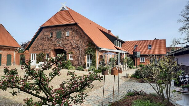 Un bâtiment traditionnel en briques, doté d’un toit à forte pente recouvert de tuiles rouges, d’une entrée en arc de pierre et d’une cour pavée agrémentée d’un banc et de plantes en pot.