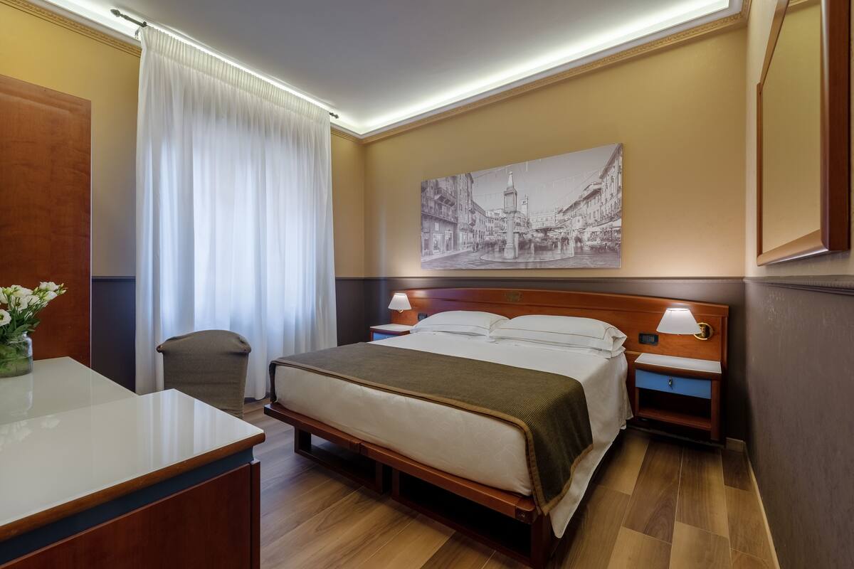 A hotel room with a bed, bedside tables, a desk, a chair, a large window with curtains, and a wall-mounted painting.