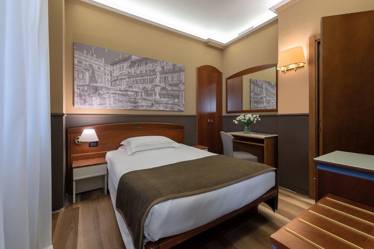 A hotel room with a bed, bedside table, lamp, mirror, and a painting of a cityscape.