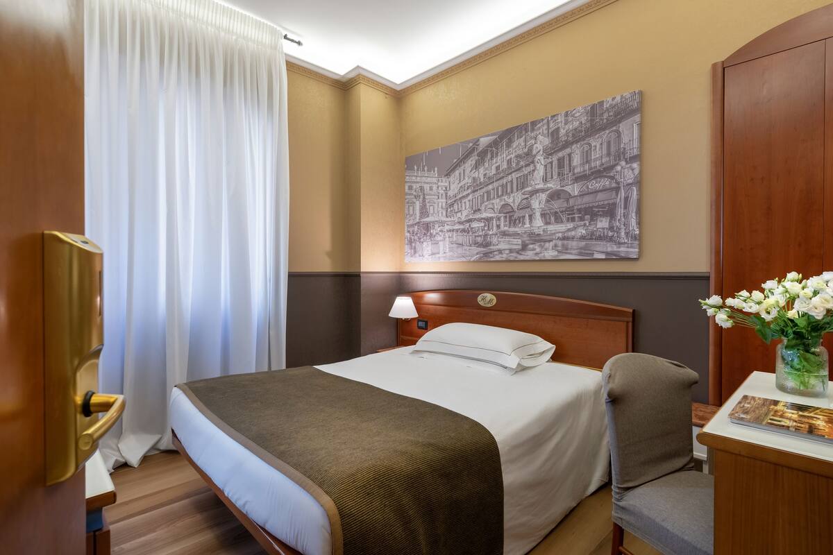 A hotel room with a bed, a nightstand, a lamp, a painting on the wall, and a window with curtains.