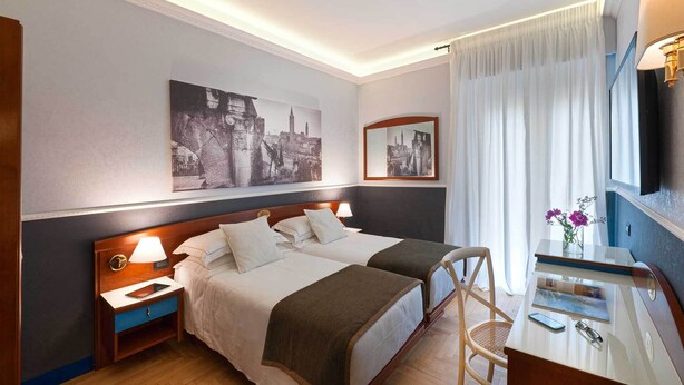 A hotel room with two beds, a desk with a chair, a nightstand, and a wall-mounted TV. - Mastino Rooms (Verona)