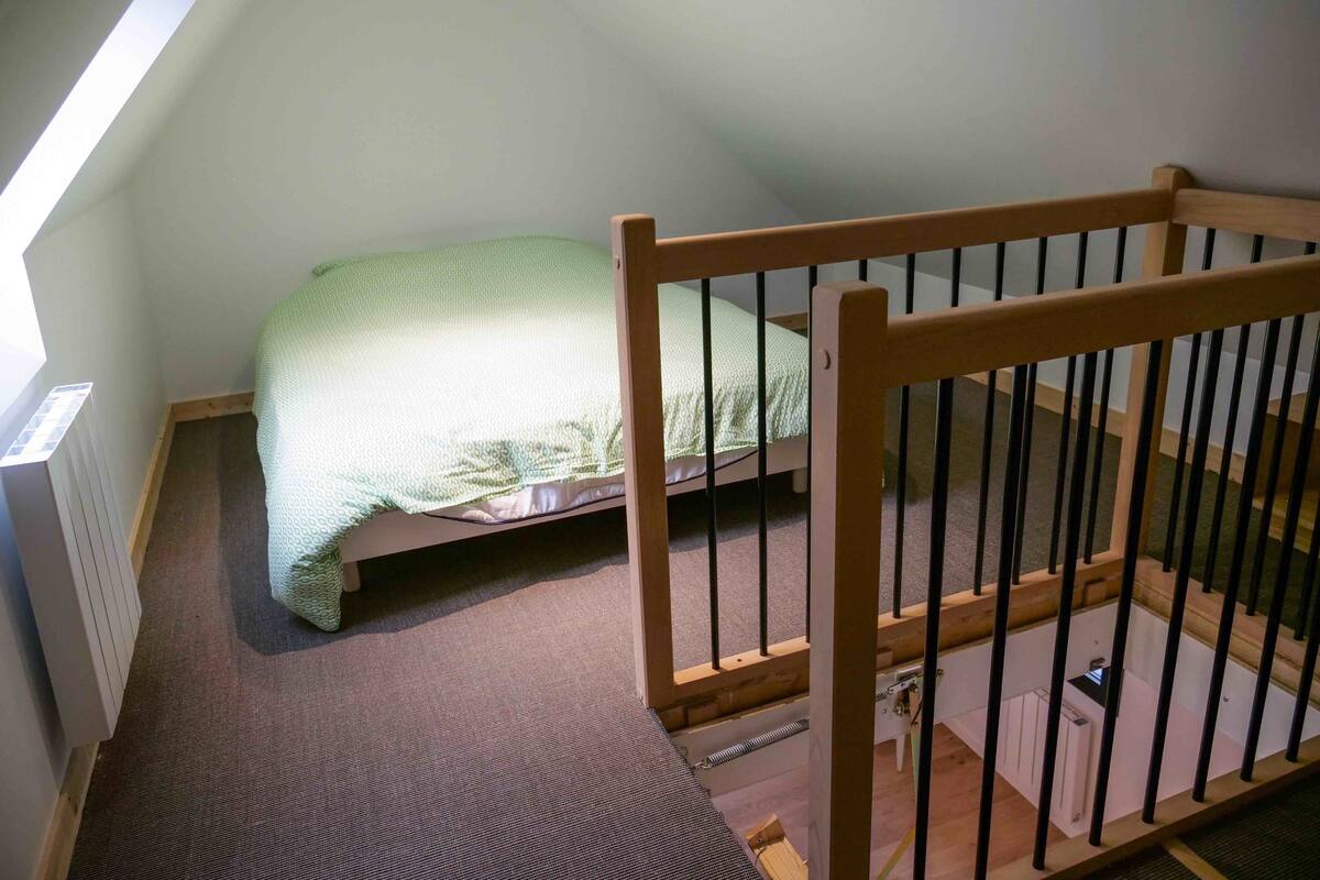 A high-ceilinged room with a sloped ceiling, a single bed with a patterned bedspread, a wooden railing, and a radiator on the wall.