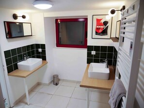 Ein Badezimmer mit zwei Waschbecken, schwarz-weißen Fliesen und einem Fenster mit rotem Rahmen, sowie einem Poster an der Wand.