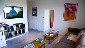 Un salon comprenant un canapé, deux tables en bois, une bibliothèque et un téléviseur fixé au mur.