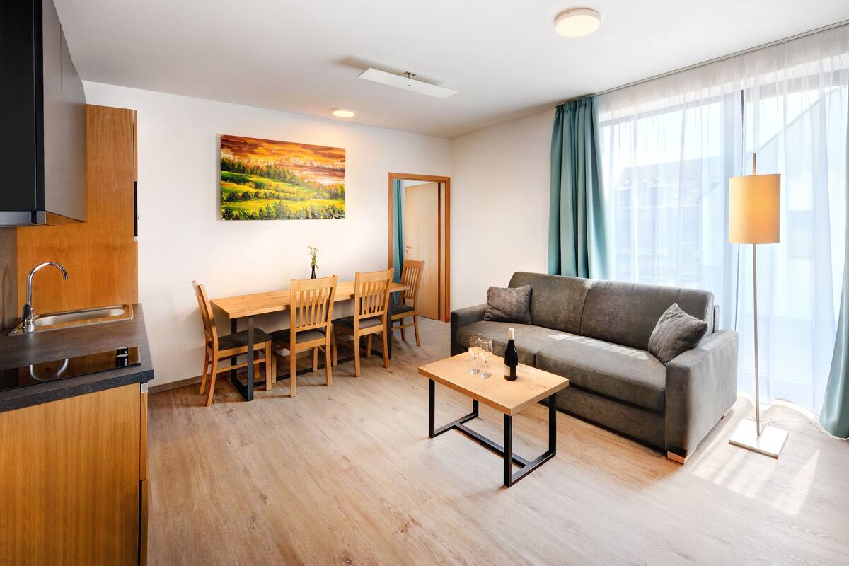A modern living room with a grey sofa, wooden dining table, and chairs. A painting is on the wall, and there's a TV on the right.