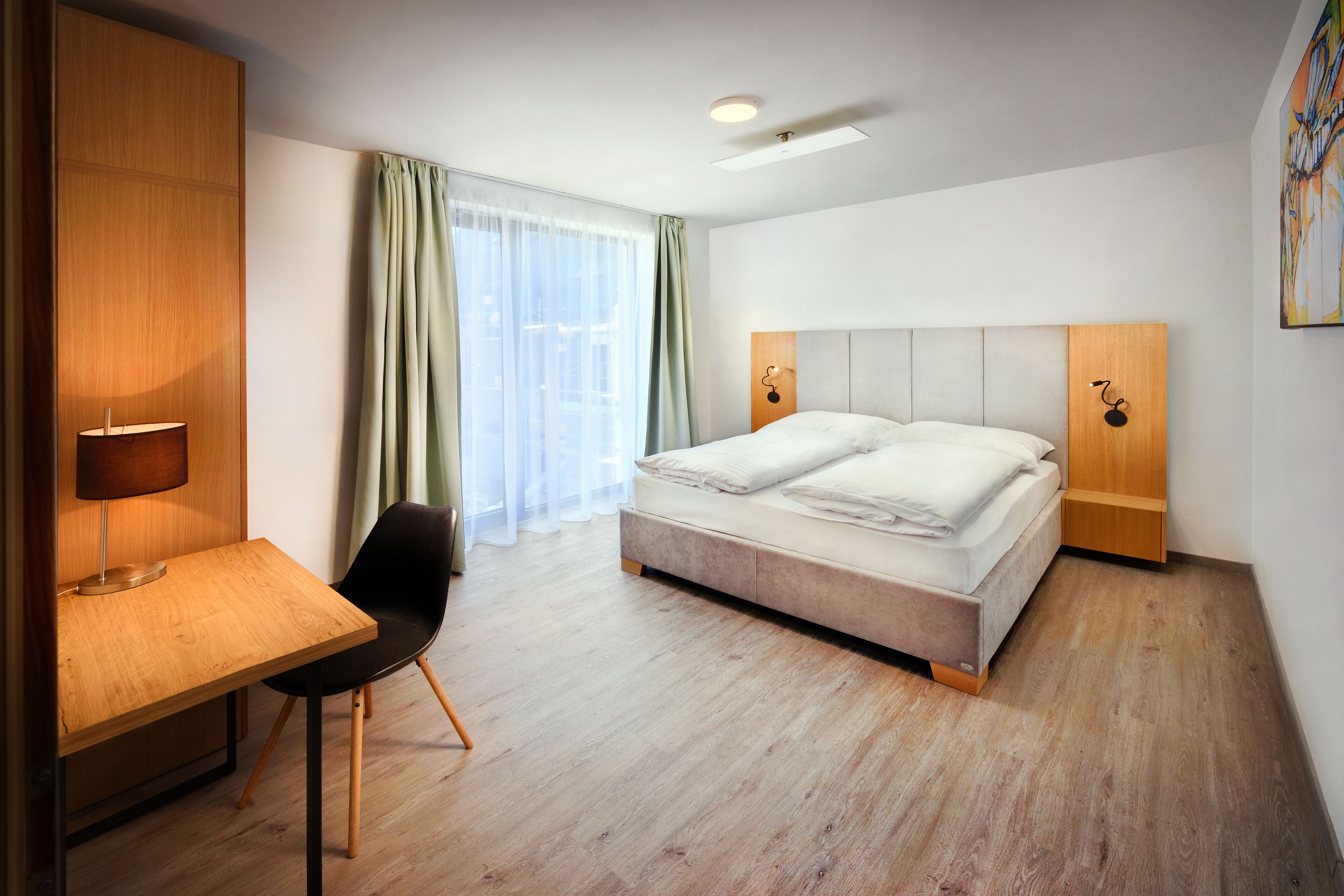 A hotel room with a bed, a desk, a chair, a lamp, and a window with curtains.