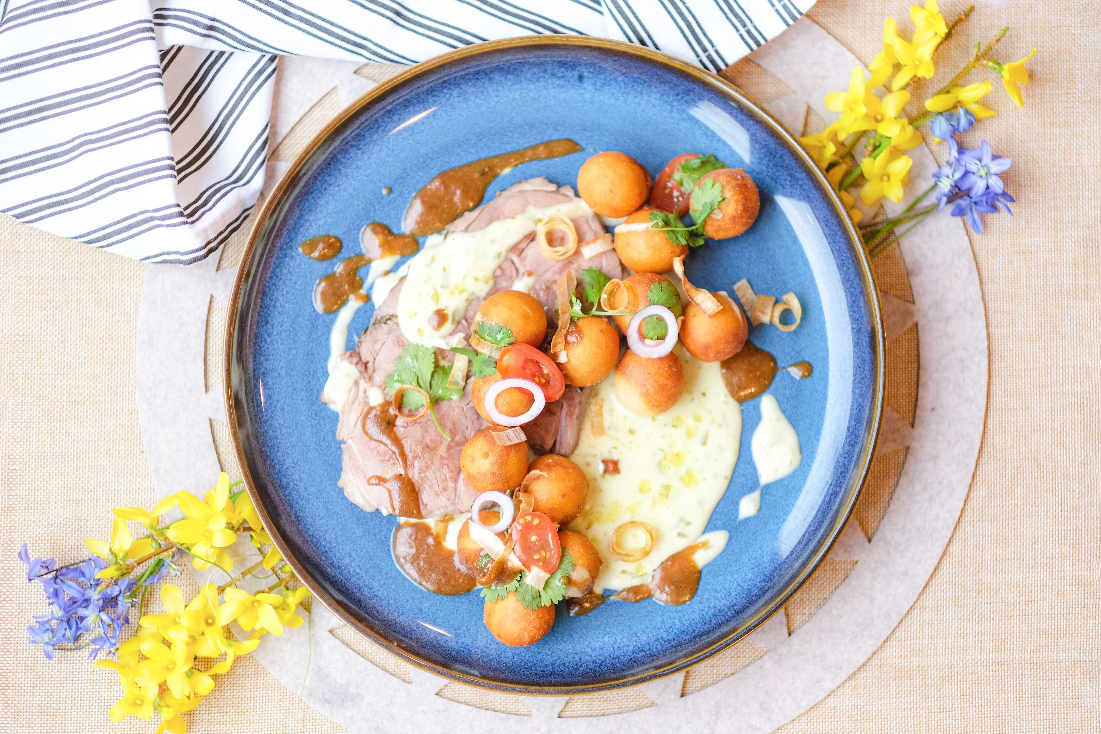 Un plato azul con comida, que incluye carne, verduras y salsa, sobre una mesa con mantel a rayas y flores.