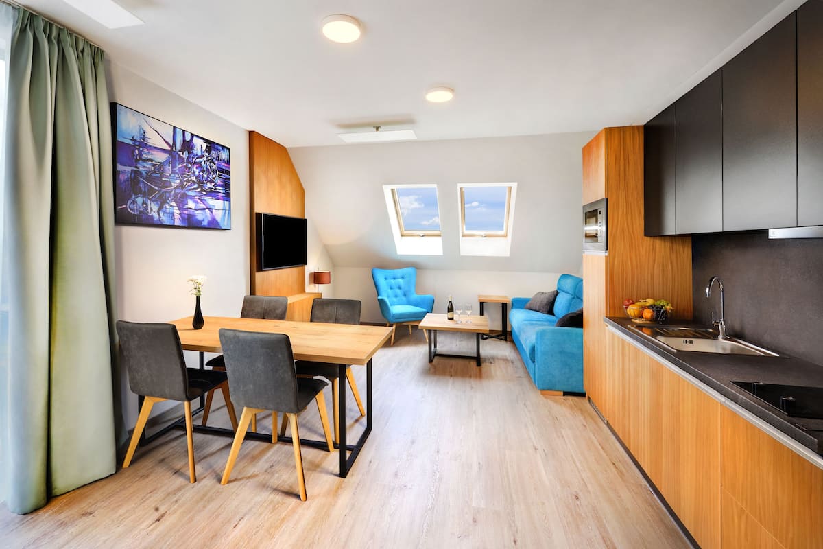 A modern apartment with a dining area, a living room with a blue sofa, a kitchen with a sink and stove, and a large window.