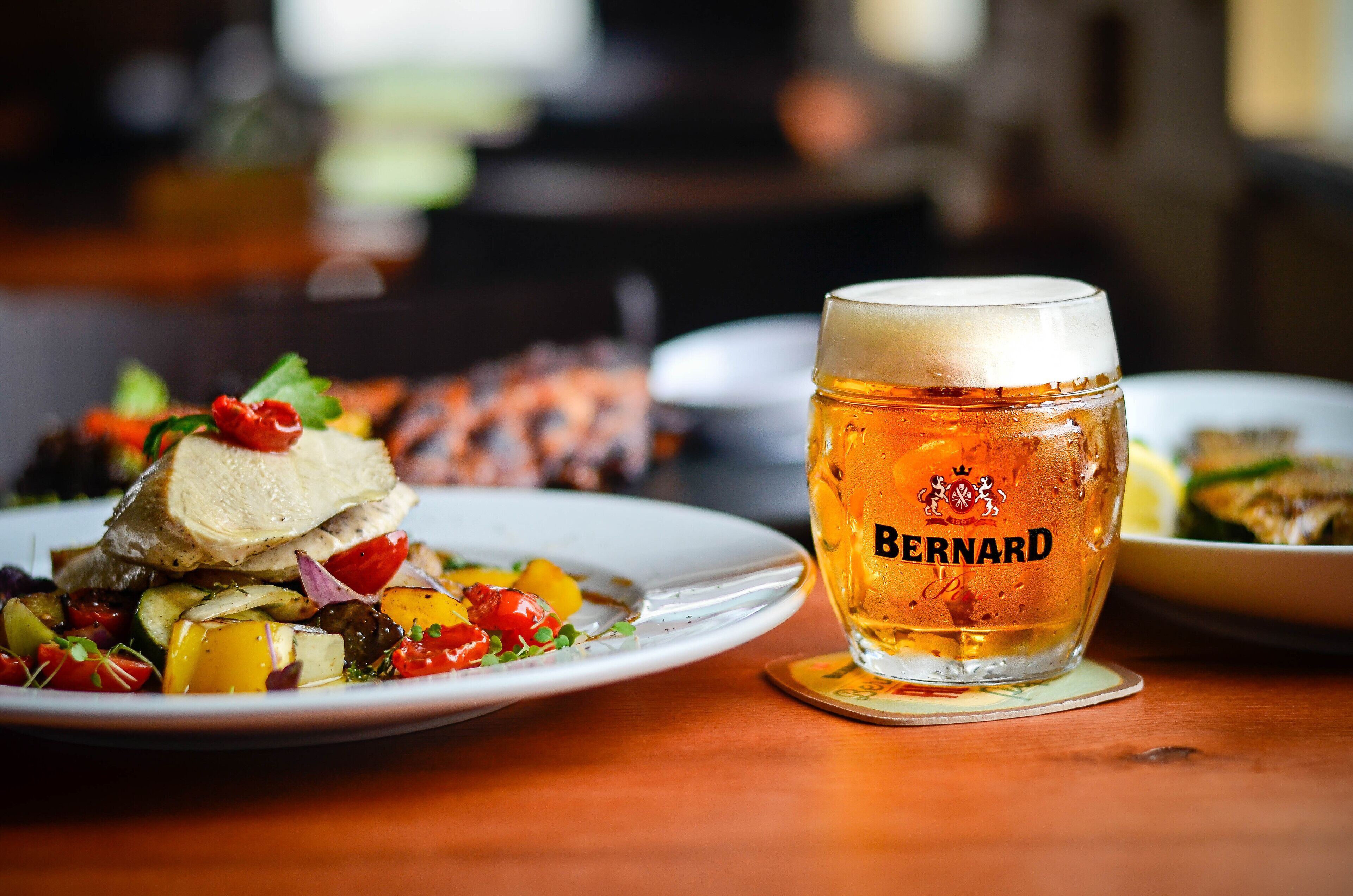 A glass of beer with the brand name "Bernard" on a coaster, a plate of food with vegetables and a piece of meat, and a blurred background suggesting a dining setting.