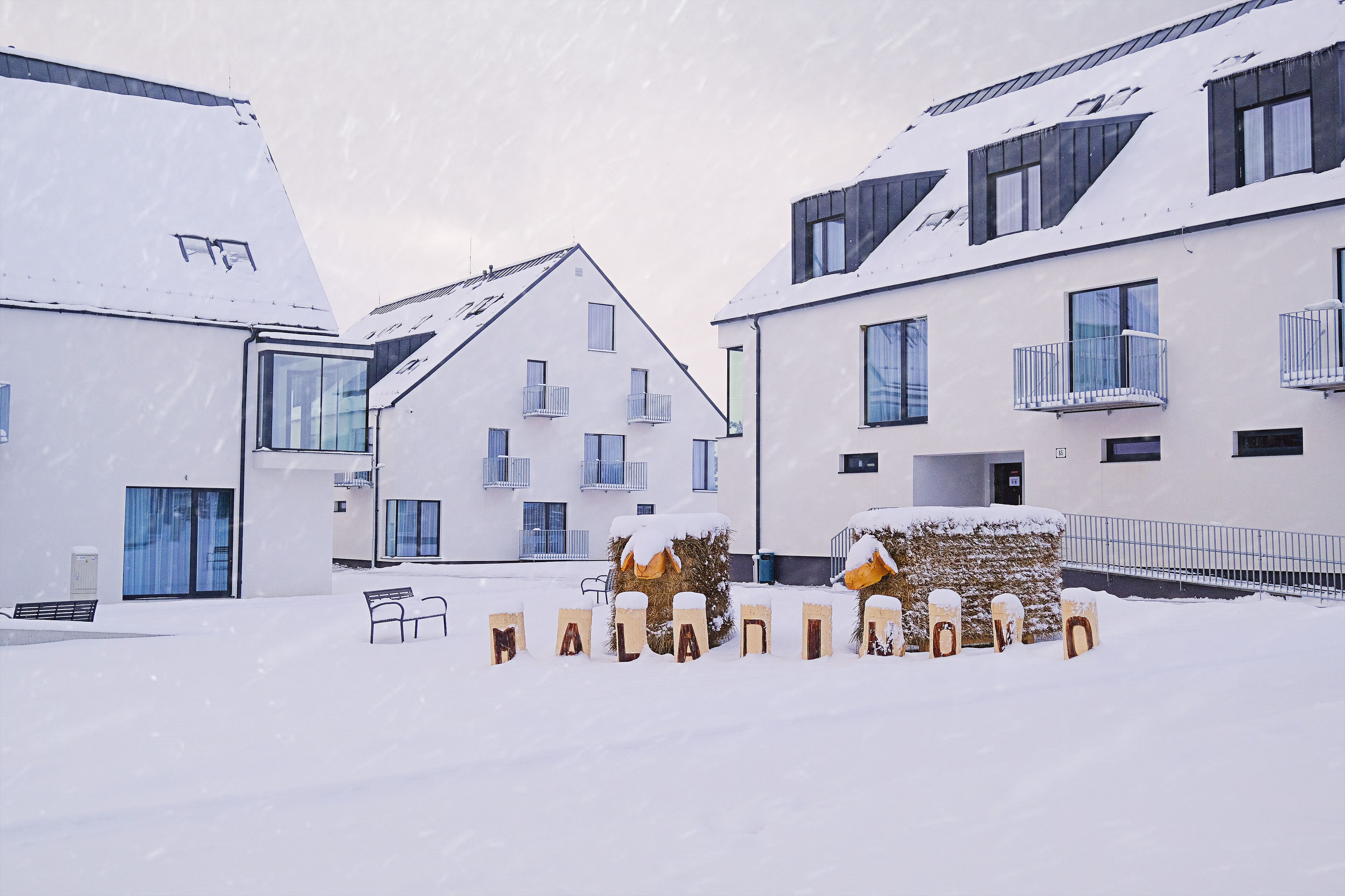 Een sneeuwbedekte binnenplaats met moderne gebouwen, een houten bank en een hooi sculptuur.