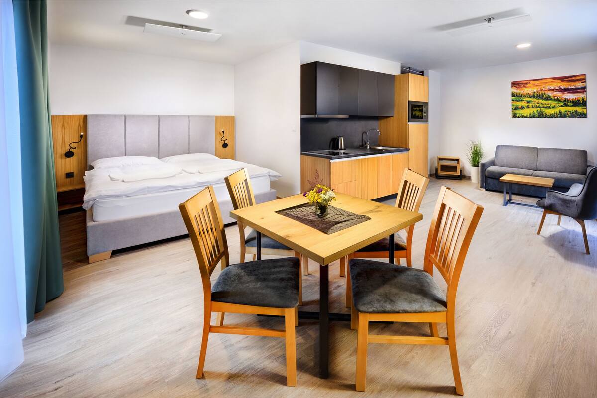 A modern hotel room with a bed, a dining area with a table and chairs, a kitchenette, a sofa, and a TV.