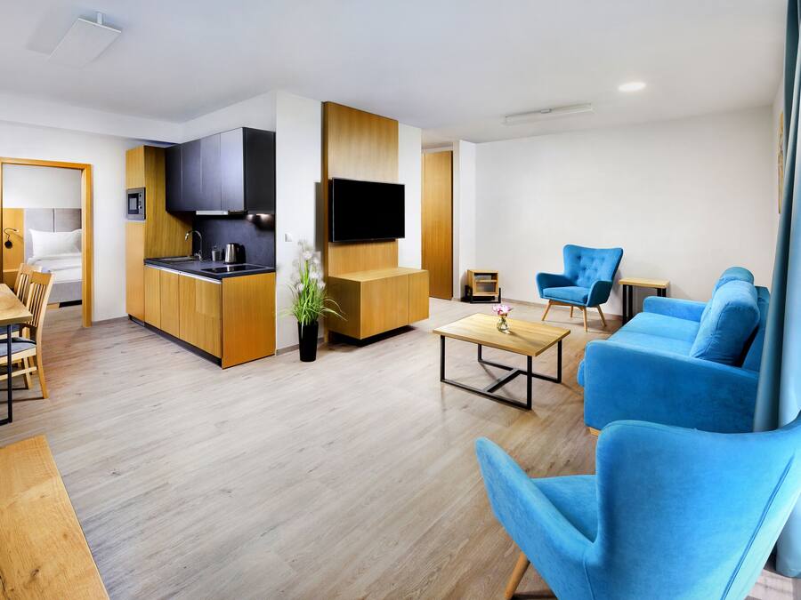 Una sala moderna con una mesa de comedor de madera, sillones azules, un sofá y una zona de cocina con un televisor.