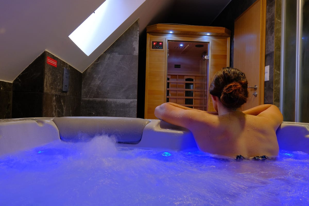 A person relaxing in a hot tub with blue lighting, a skylight, and a wooden door.