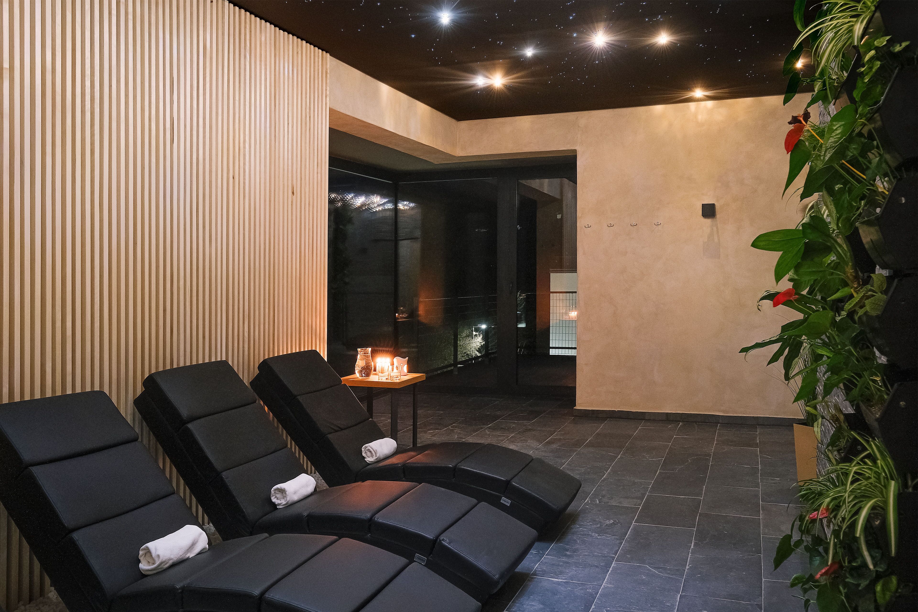 Un área de spa con sillones reclinables negros, una mesita con velas y una pared cubierta de plantas.