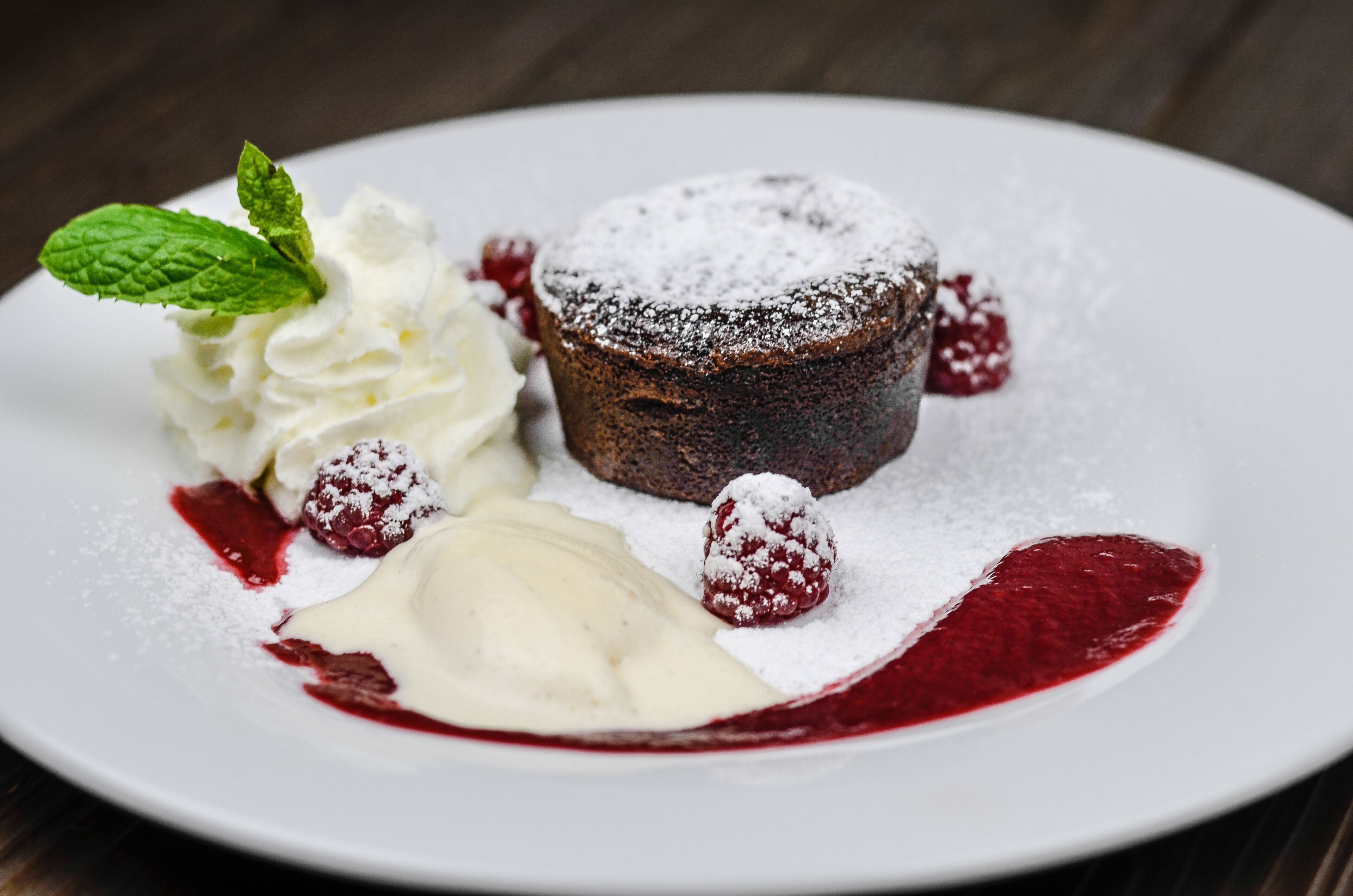 Un plato de postre con pastel de chocolate, crema batida, frambuesas y una hoja de menta.