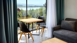 A modern living room with a wooden table and two black chairs, a grey sofa, and a large window offering a view of a lake and greenery. - Maladinovo (Liptovsky Mikulas)
