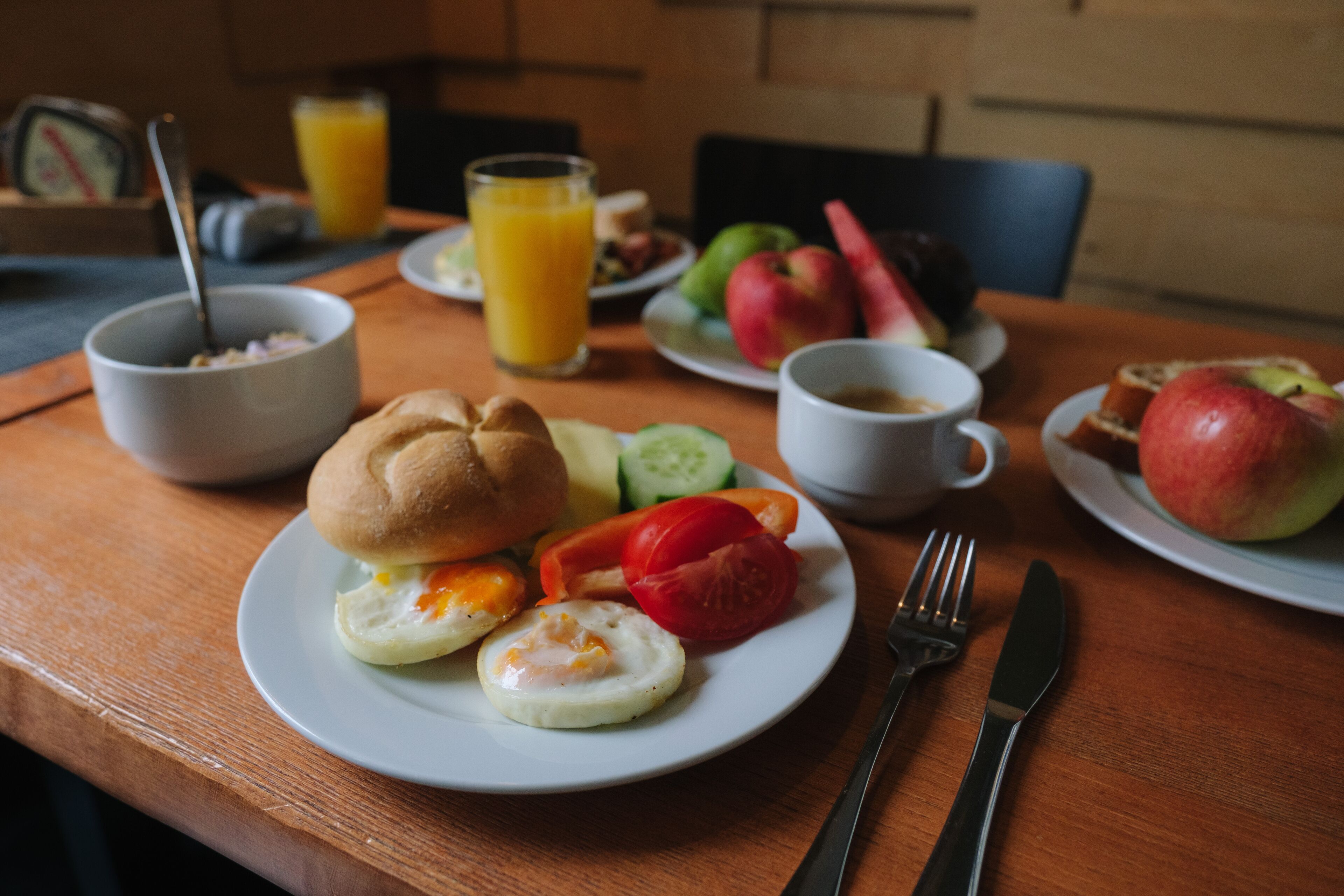 A breakfast table with a plate of eggs, vegetables, and a sandwich. There are also a glass of orange juice, a cup of coffee, and a plate with fruits.
