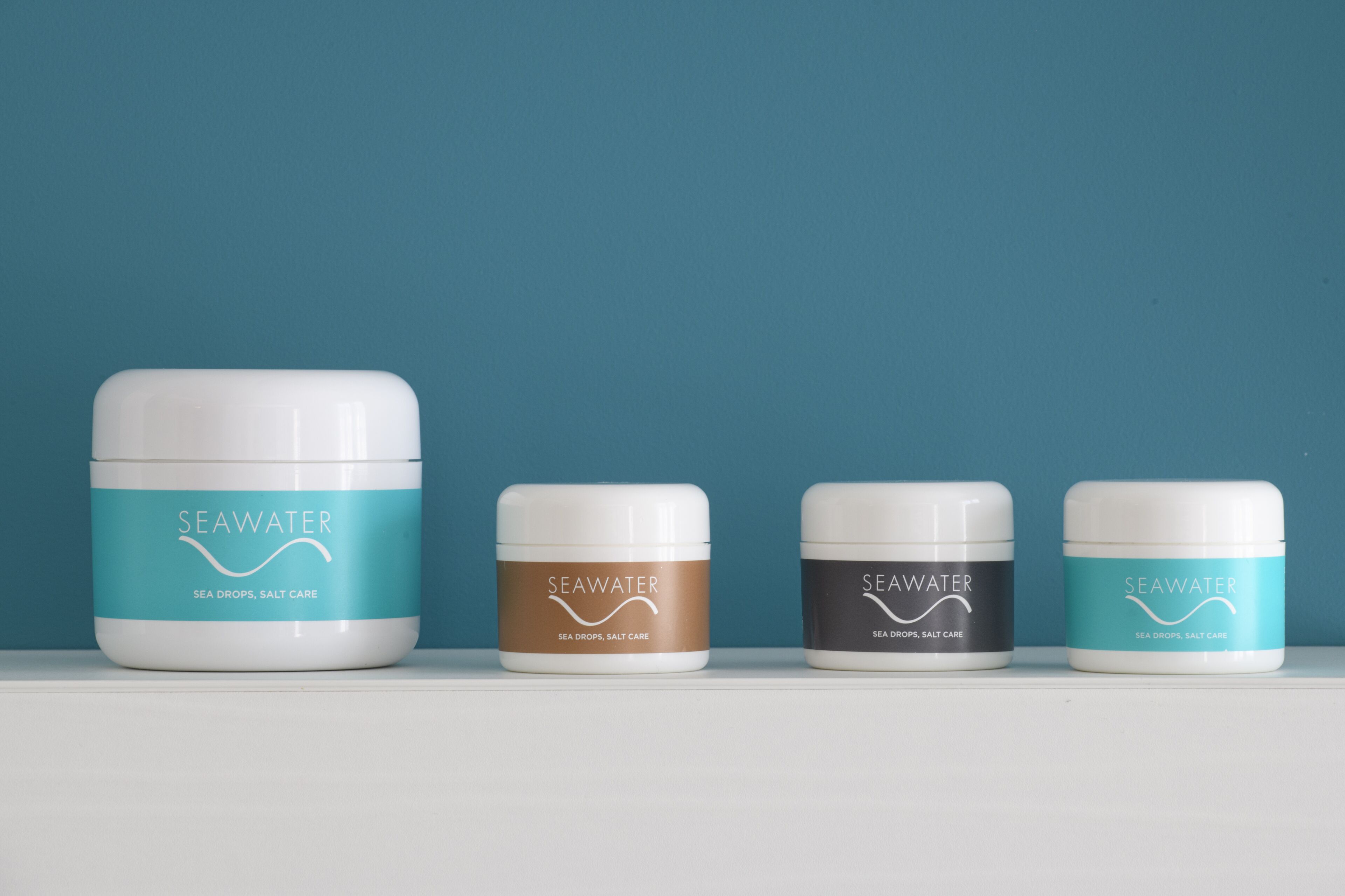 Vier potjes SeaWater sea drops, verzorging met zout op een witte plank voor een blauwe achtergrond.