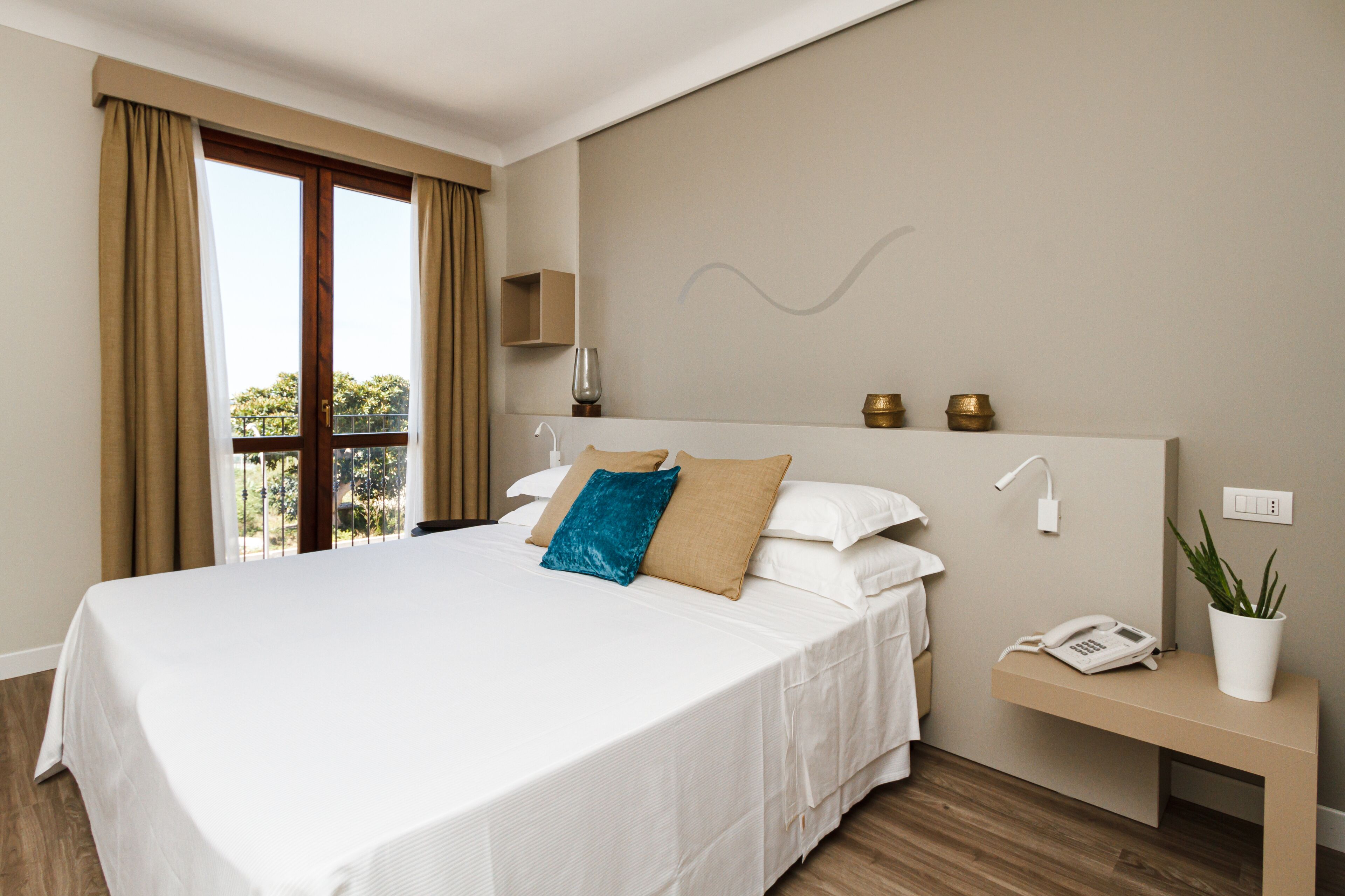 A hotel room with a large bed, a bedside table with a phone and a plant, and a view of the outdoors through a window.