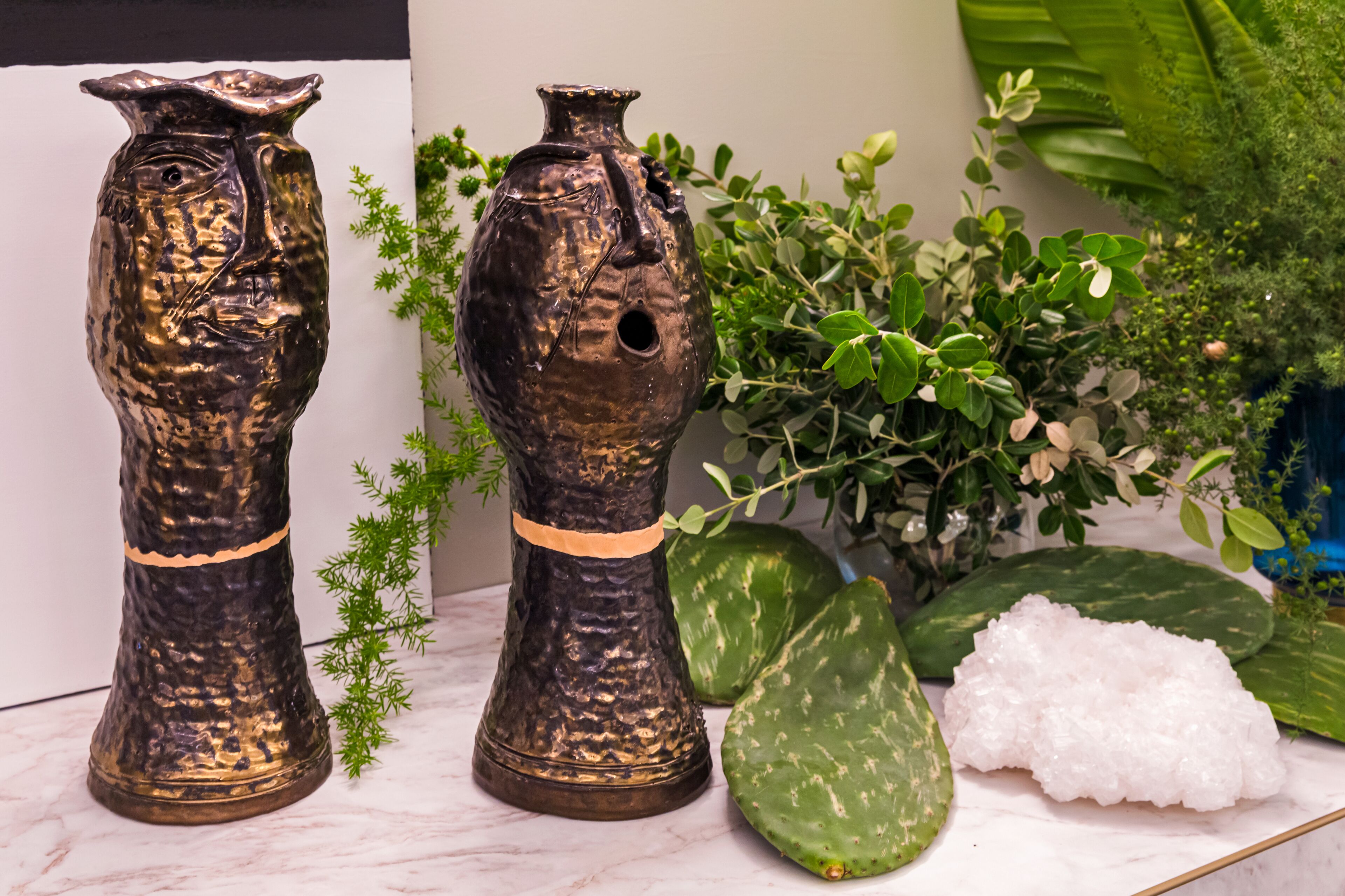 Twee metalen vazen met een geribbelde oppervlakte en een klein gaatje in de rechtervaas, staan op een marmeren oppervlak met groene planten en een witte kristal erbij.
