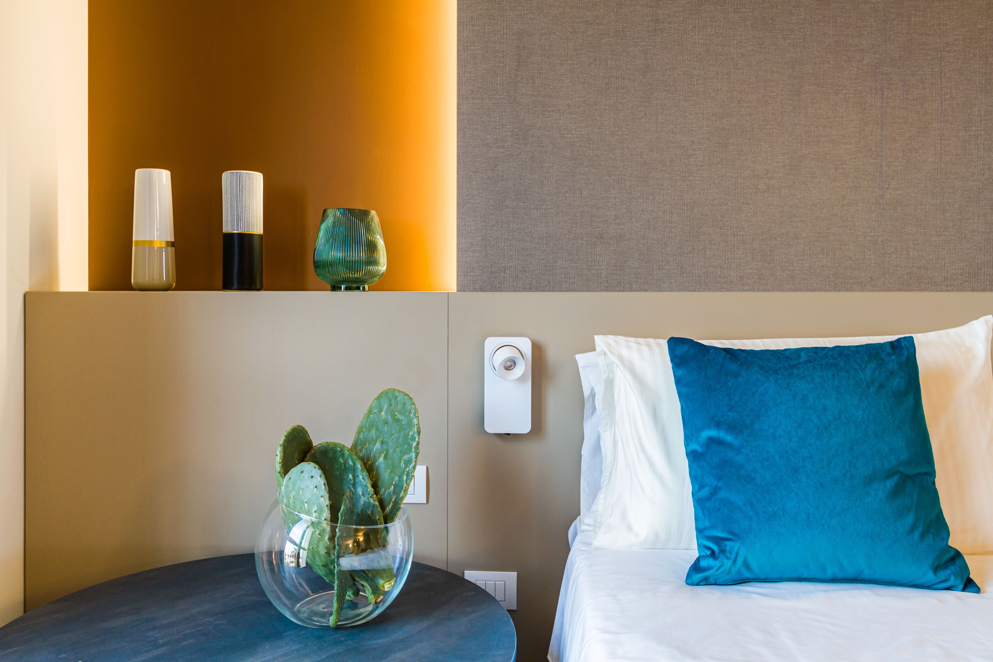 A hotel room with a bed, a nightstand with a vase of cactus, a wall-mounted alarm clock, and a shelf with two bottles.