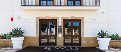 A hotel entrance with a double door, a balcony with a railing, and potted plants on either side.