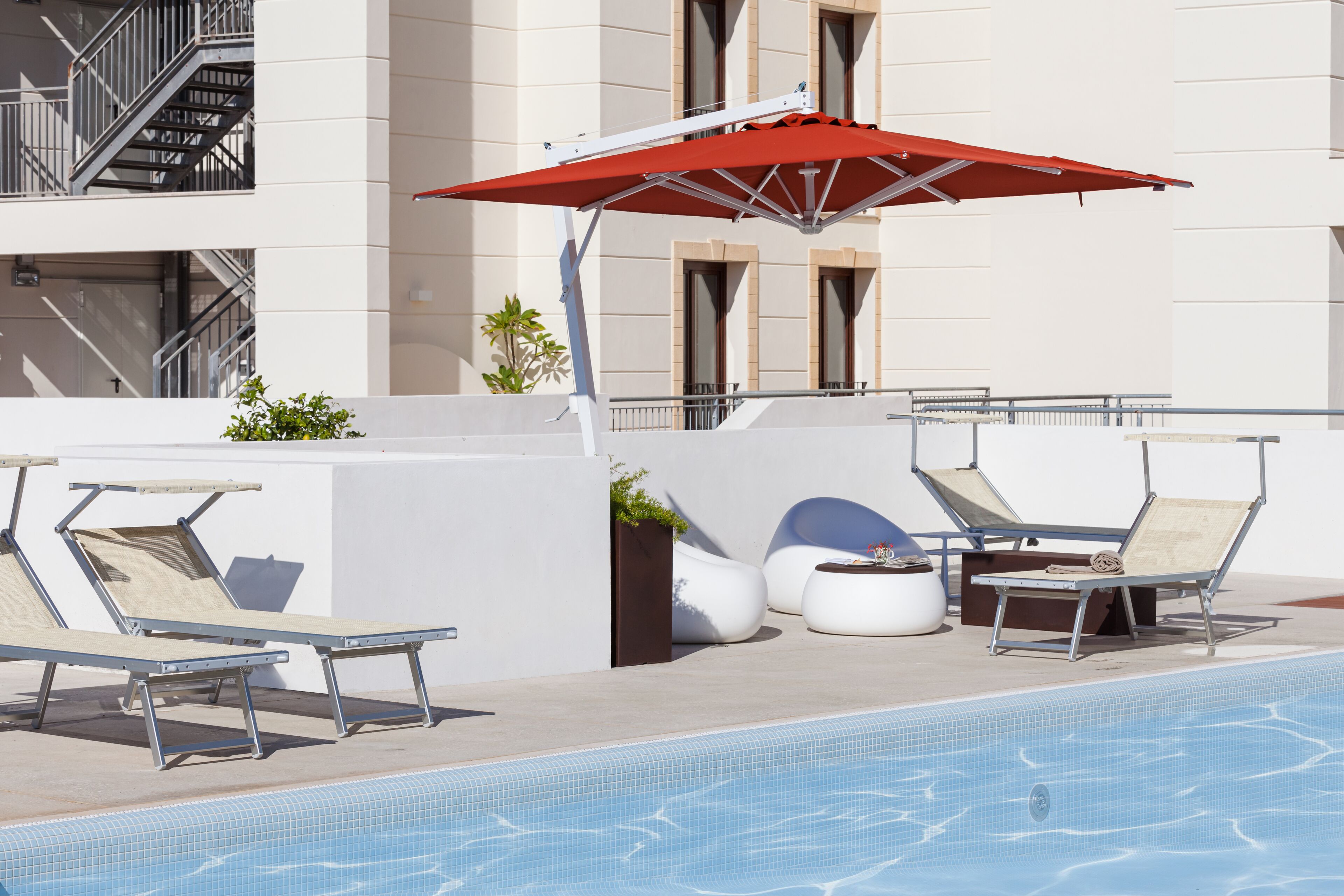 A poolside area with lounge chairs, a red umbrella, and a modern building in the background.