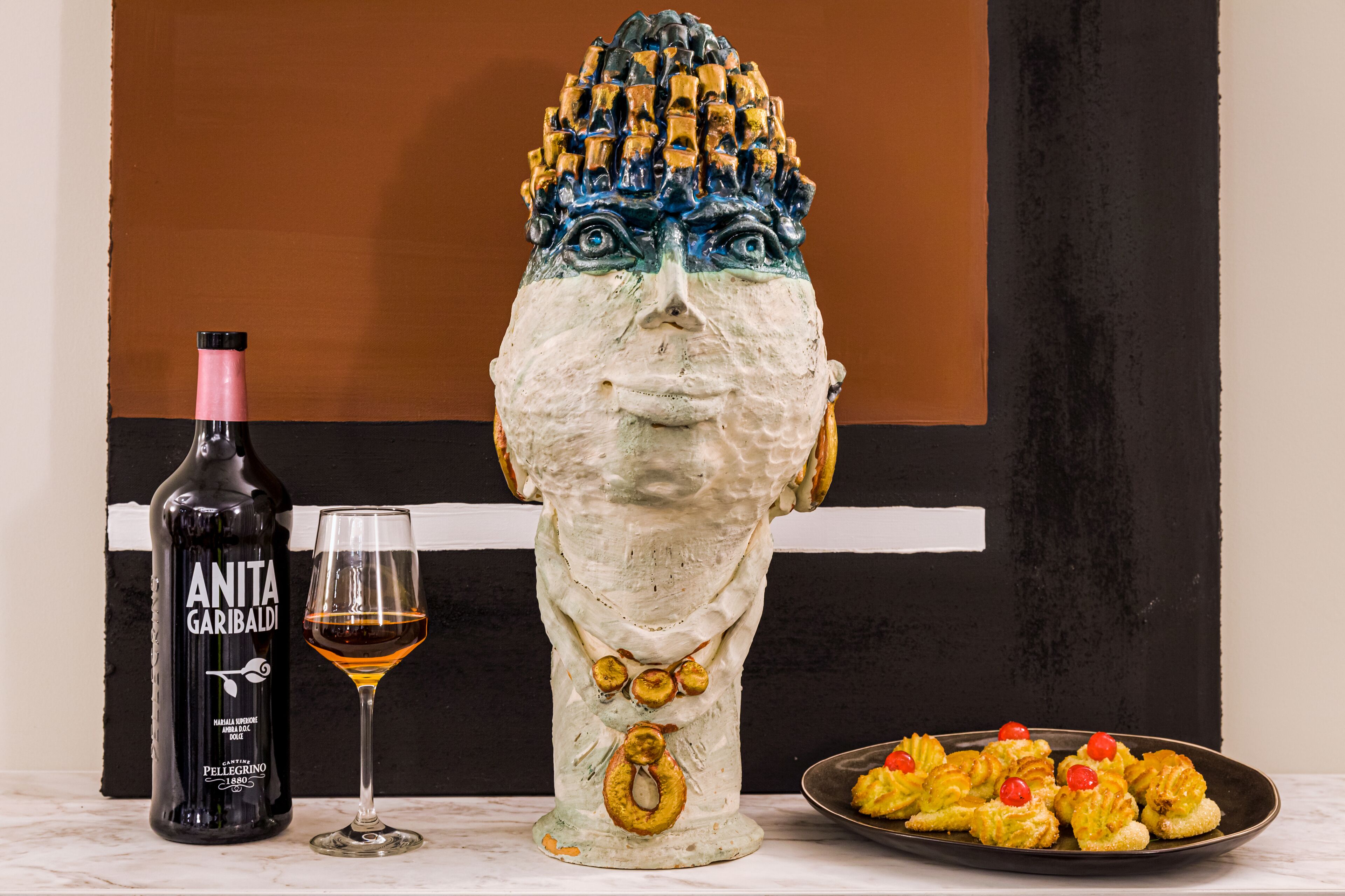 Une sculpture représentant un visage, avec une bouteille de vin Anita Garibaldi et un verre à côté, et une assiette de nourriture au premier plan.