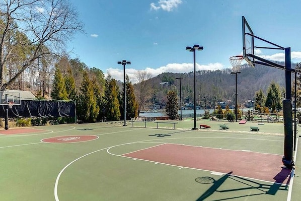 An outdoor basketball court with a green and red playing surface, surrounded by trees and a mountain in the background.