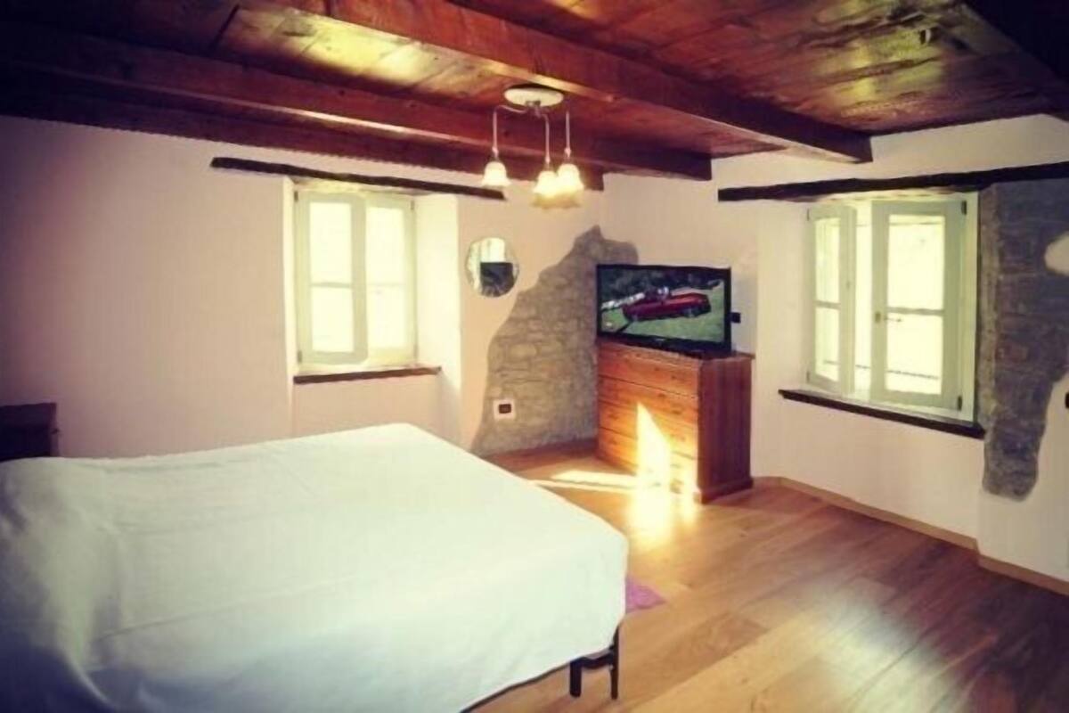 Un dormitorio con cama, televisión, paredes de piedra, vigas de madera y ventanas amplias.