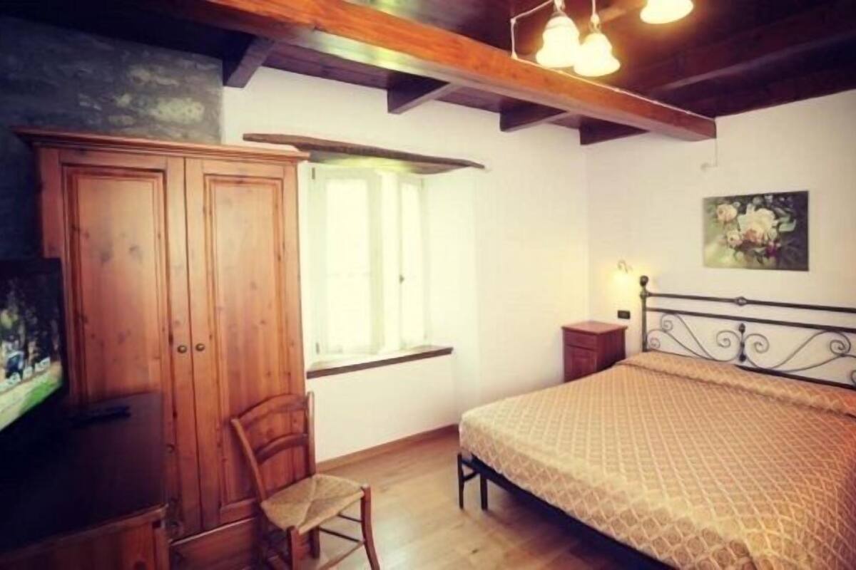 Un dormitorio con un armario de madera, una cama con colcha estampada, una silla, un ventanal con cortinas y un cuadro en la pared.