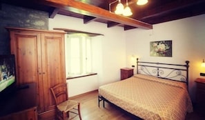 A bedroom with a wooden wardrobe, a bed with a patterned bedspread, a chair, a window with curtains, and a painting on the wall. - Agriturismo La Luna (Riolunato)
