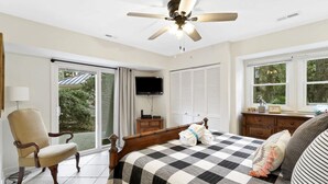 Un dormitorio con cama, una silla, televisor, ventilador de techo y ventana.