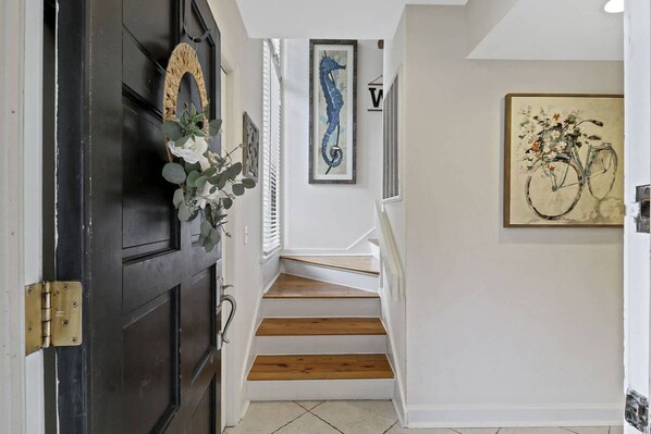 A hallway with a black door, a staircase with wooden steps, and wall-mounted artwork including a seahorse and a bicycle painting.